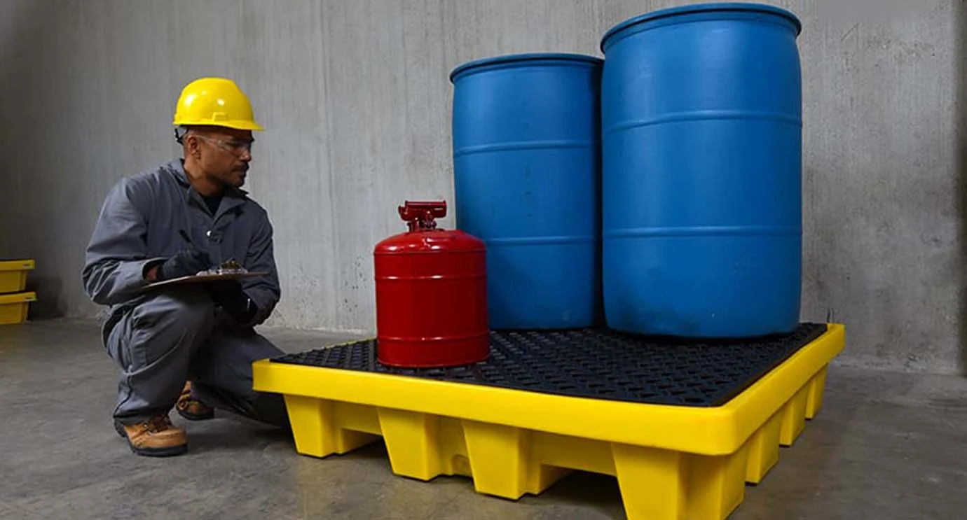 Industrial worker inspecting chemical drums stored on yellow spill pallet for leak prevention and safe storage